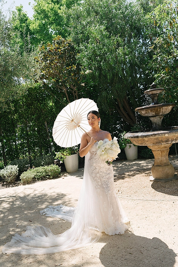 Bridal portrait of a bride in a strapless lace gown with long train, holding a white bouquet and parasol by a stone fountain courtyard