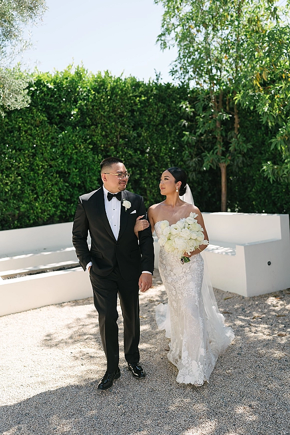 Couple portrait of bride in strapless lace dress and veil holding a white rose bouquet beside groom in black tuxedo with glasses in a courtyard
