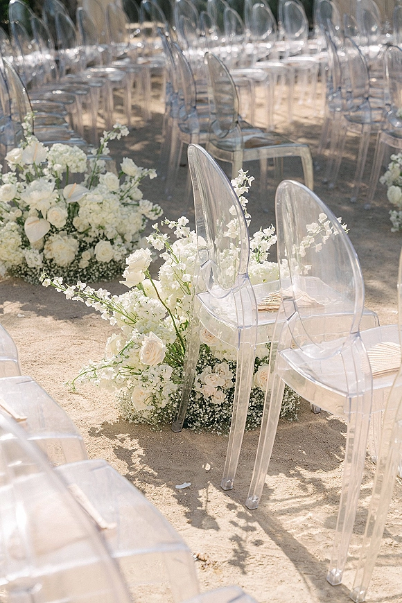Ceremony aisle decor with outdoor ceremony seating, clear acrylic chairs and white rose and baby’s breath florals lining a sand aisle