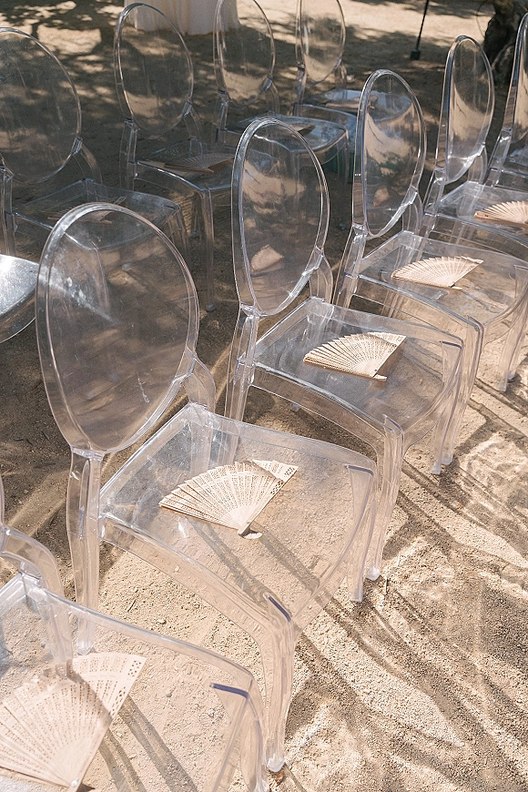 Ceremony seating with clear acrylic wedding chairs lined in rows, each with a hand fan on the seat, set on outdoor ground with tree shadows