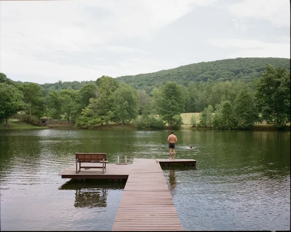 Lakeside dock and wooden lake pier with a simple bench, stretching over calm water toward wooded hills beneath a soft, cloudy sky.