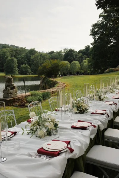 Outdoor reception tablescape featuring garden reception decor and burgundy napkins, set on a lakeside lawn amid trees and a stone wall.