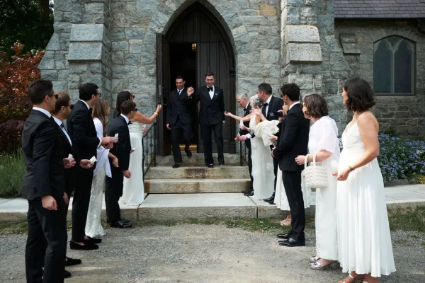 Ceremony exit with a joyful bubble send off as the couple in tuxedos walks down the stone church steps beneath an arched wooden door.