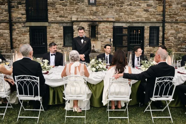 Wedding toast with the groom giving a speech at an outdoor table, taper candles aglow against a stone wall with black windows on the lawn.