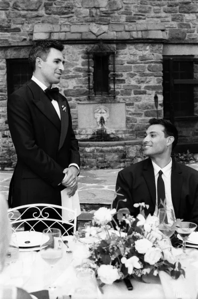 Wedding speech and reception toast by a guest in a bow tie, guests listening around dinner tables in a stone-walled courtyard with a fountain.