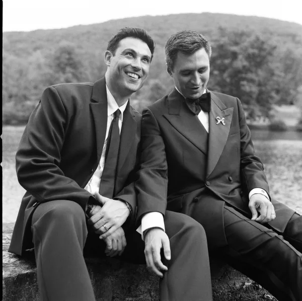 Couple portrait of two grooms sitting, laughing by a lakeside stone wall, wedding bands catching light against the trees.