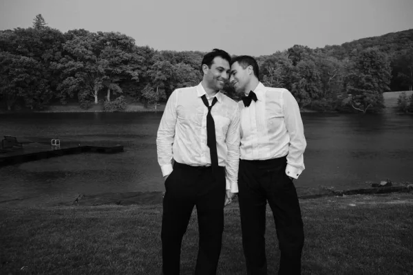 Couple portrait of two grooms sharing a gentle forehead touch on a dock by a lake, in crisp dress shirts, with trees and hills beyond.