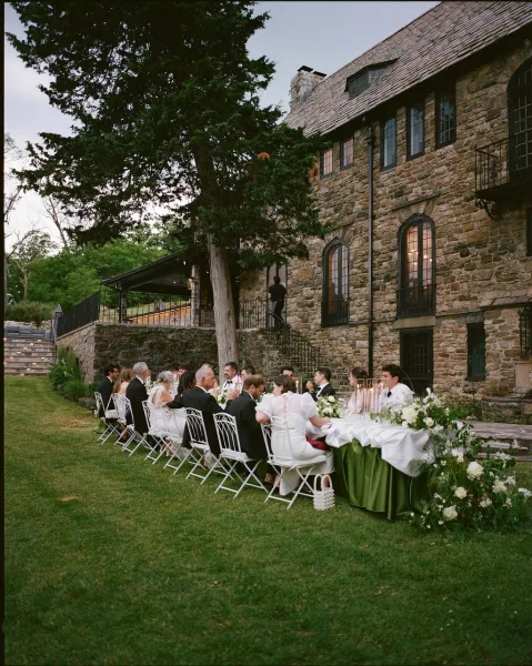 Outdoor reception dinner set as an al fresco wedding reception along a long banquet table beside a stone building on a tree-lined lawn