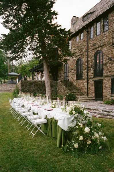 Reception tablescape for an outdoor reception table with taper candles on a lawn before a stone facade and trees.