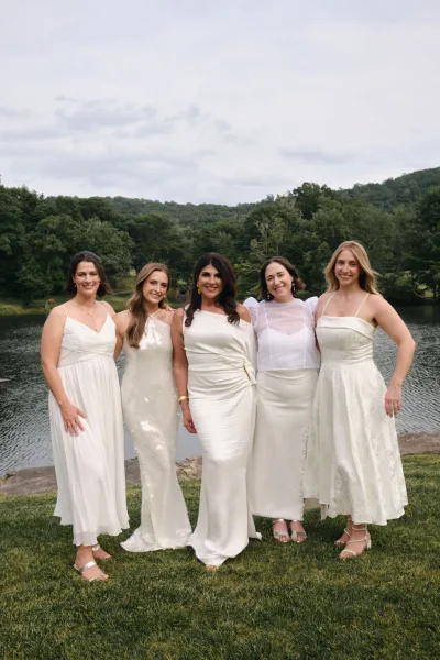Bridal party portrait of a wedding party in white, standing in strappy heels by a lakeside lawn with trees and hills under a softly cloudy sky.