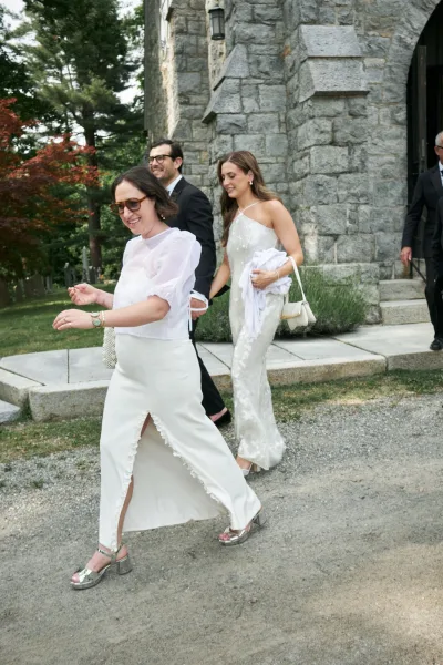 Wedding guests spill out in a joyful church guest exit by a stone church exterior, one wearing a chic one-shoulder dress on the pathway.