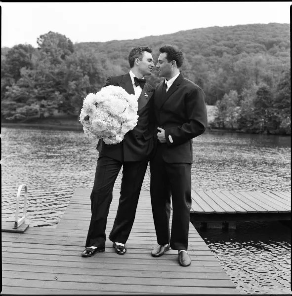 Couple portrait of two grooms, one holding a bouquet, standing on a wooden dock by a calm lake with forested hills rising in the background.