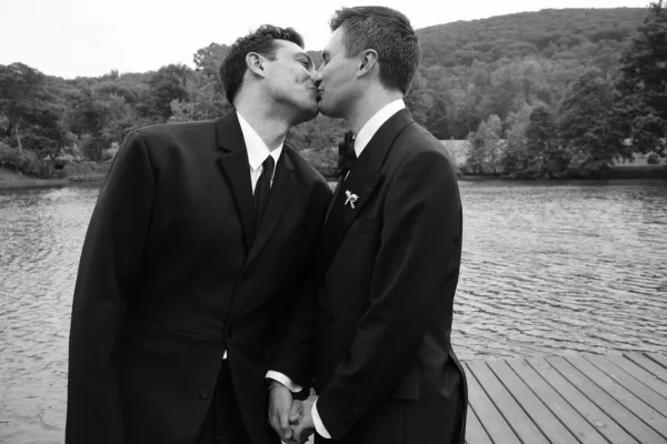 Wedding kiss portrait as groom and groom kiss on a wooden dock by a calm lake, wearing tuxedos, with trees and hills beyond.