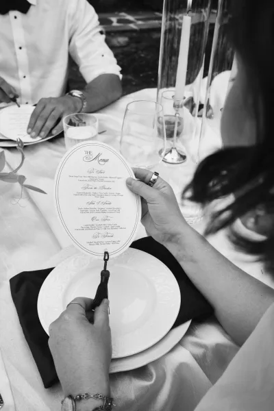 Wedding menu on an oval menu card with a tassel rests on a tablecloth-covered tabletop, patio stones softly visible in the background.