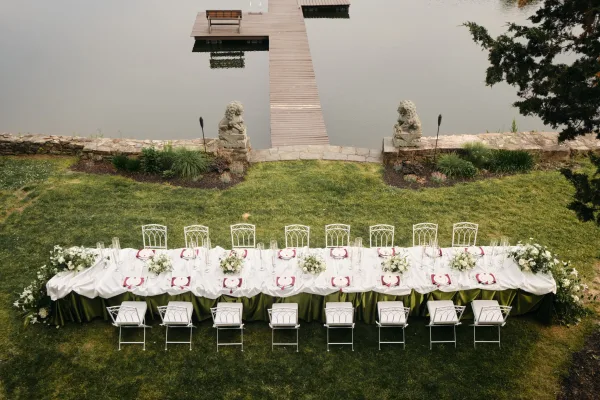 Reception tablescape on a long banquet table with glass hurricanes, set on a lakeside lawn beside a wooden dock and stone wall.