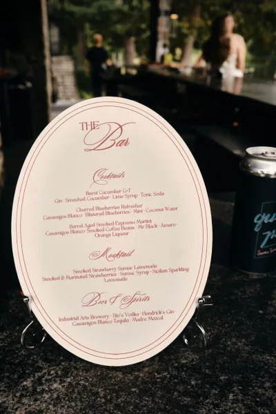 Bar menu sign with a wedding bar menu printed on an oval card, set on a bar counter with trees and a stone wall in the background.