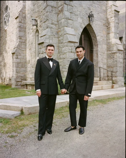Couple portrait of two grooms holding hands in black suits, a bow tie accent, standing by an arched stone church doorway on the steps.