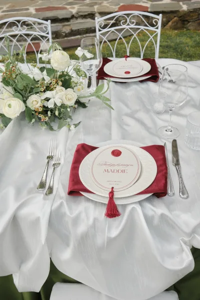 Wedding place setting featuring refined reception table decor and a burgundy napkin on a garden lawn with stone steps in the background.