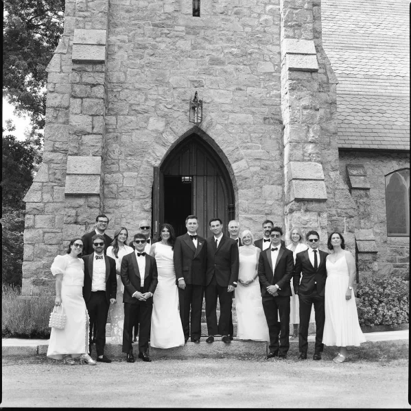 Wedding party portrait on the stone church steps beneath an arched doorway, a cheerful group photo featuring playful sunglasses.