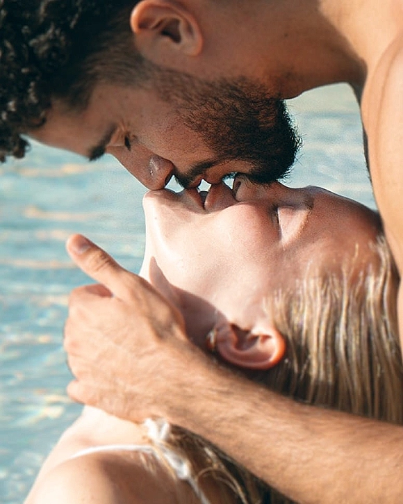 Wedding kiss as the groom kisses the bride close up, his hand on her face, with ocean water softly blurred behind them