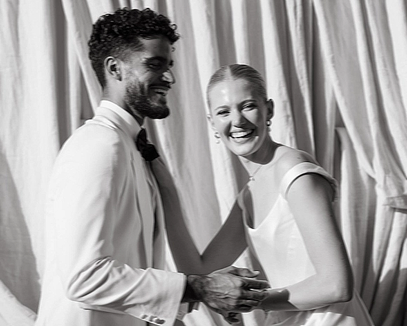 Couple portrait in a black and white wedding portrait style, bride in sleek dress and groom in white suit laughing before a draped backdrop