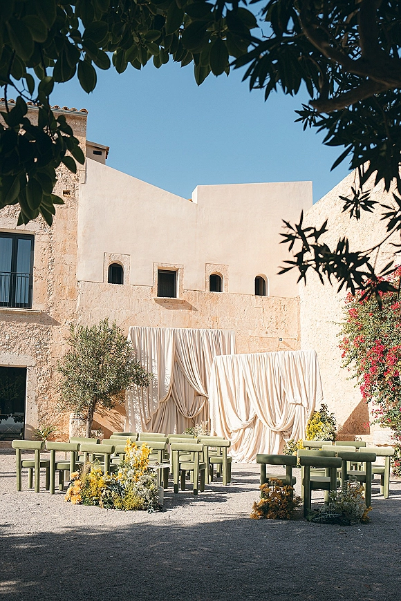 Ceremony setup with outdoor ceremony seating facing a draped fabric backdrop, with white and yellow florals in a stone courtyard under blue sky