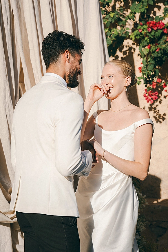 Wedding couple portrait of bride and groom laughing while holding hands, bride in off-shoulder satin dress before sunlit draped stucco wall