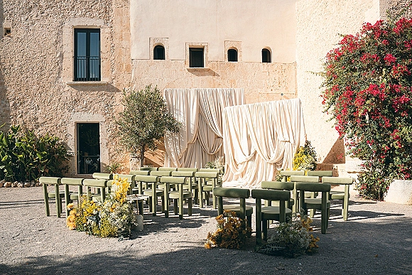 Ceremony setup with outdoor ceremony seating on green benches facing a draped fabric backdrop, with yellow aisle flowers in a stone courtyard