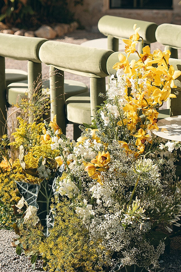 Ceremony aisle flowers in yellow orchids and white blooms with baby’s breath and greenery, arranged on gravel beside green chairs outdoors