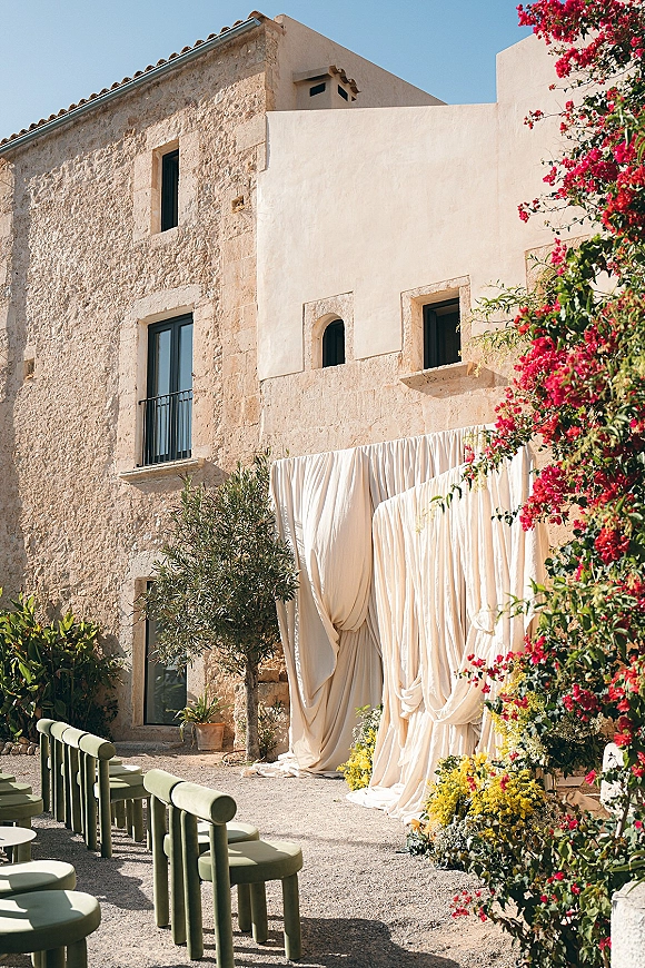 Ceremony setup with outdoor wedding ceremony setup draped fabric backdrop, flower arrangements, olive tree, and chairs in a stone courtyard