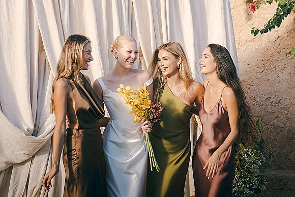 Bridesmaid group portrait with bride with bridesmaids laughing arm in arm, holding bouquets in mismatched satin dresses by a stucco wall