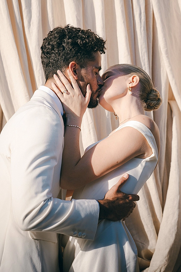 Wedding kiss portrait of bride and groom kissing close up, groom in a white suit as she touches his face, against a draped fabric backdrop
