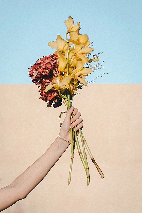 Wedding bouquet with yellow orchids and burgundy hydrangea stems held up against a blue sky and beige wall backdrop