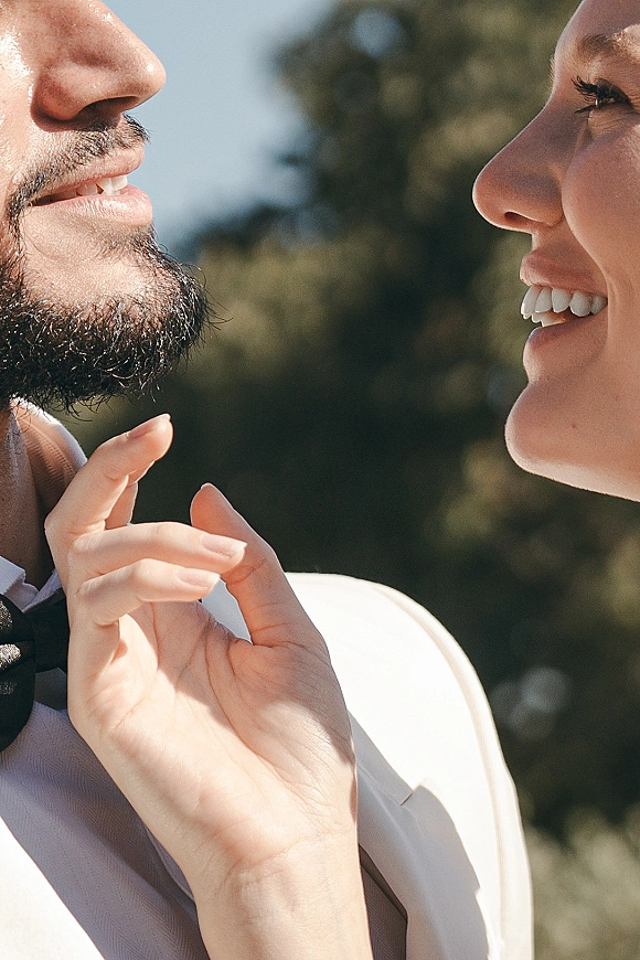 Couple portrait of bride touching groom’s face forehead to forehead, groom in light suit jacket and black bow tie against greenery and blue sky