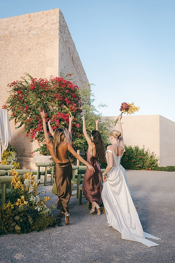 Bridesmaids with bride walk away in colorful dresses, bride lifting bouquet overhead in a stone courtyard with flowering vine wall﻿