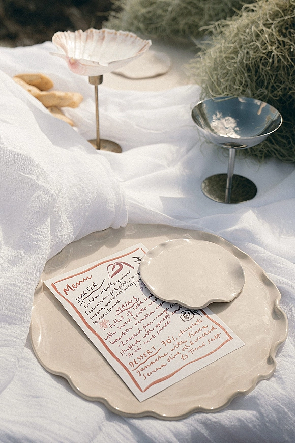 Wedding tablescape with a handwritten wedding menu beside scalloped ceramic plates, coupe glasses, bread rolls, and dried moss in outdoor light