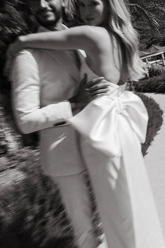 Wedding couple portrait in a black and white wedding photo, bride in strapless gown hugging groom in light suit with boutonniere in a garden lawn by a house exterior