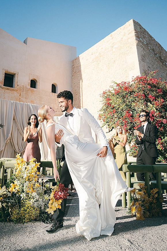 Wedding couple portrait of groom dipping bride as she holds an orchid bouquet, veil flowing in a sunny stone courtyard with vine blooms