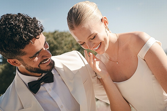 Couple portrait of bride and groom laughing, bride leaning on him in off-shoulder dress and bow tie suit under blue sky and trees