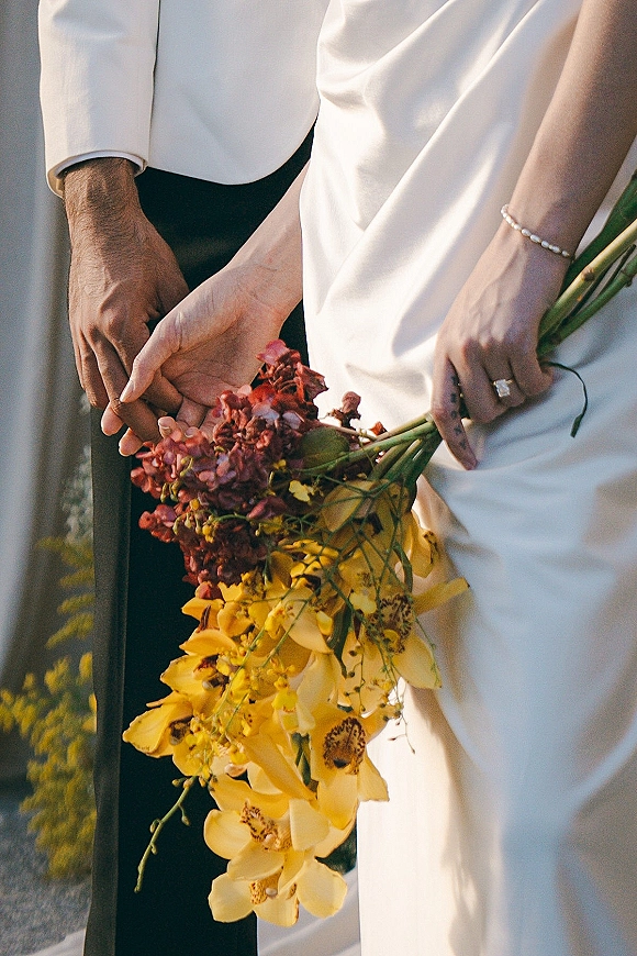 Couple holding hands, wedding hands close up with rings as bride holds yellow orchid and burgundy bouquet on a sunlit garden path