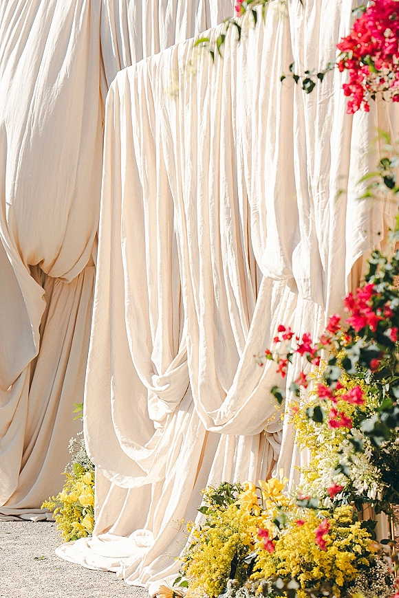 Ceremony backdrop with draped fabric and pink-and-yellow florals, greenery at the base, set along a sunlit outdoor walkway