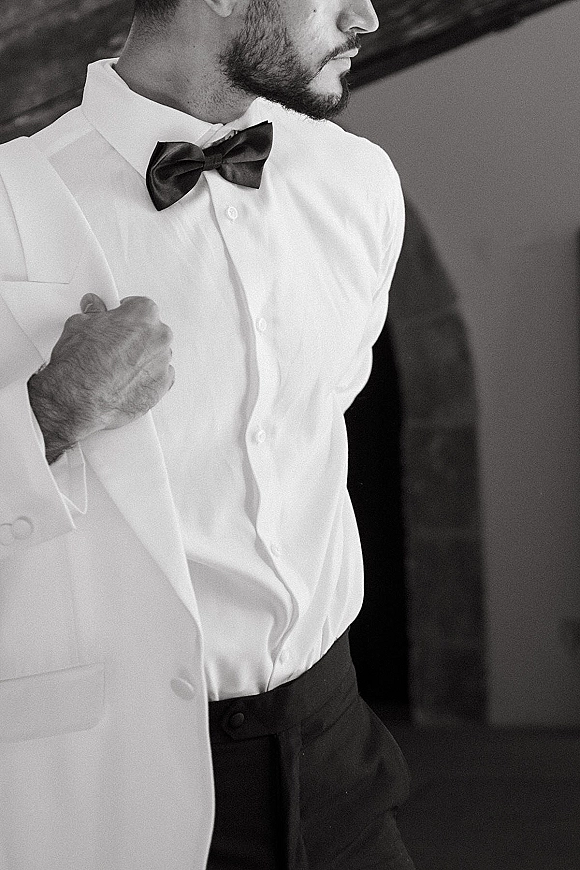 Groom portrait in a white dinner jacket with black bow tie and suspenders, standing by an indoor doorway shadow in formal attire