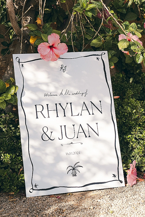 Wedding welcome sign with calligraphy lettering and hibiscus greenery accents, leaning by garden shrubs along a gravel path near a tree trunk