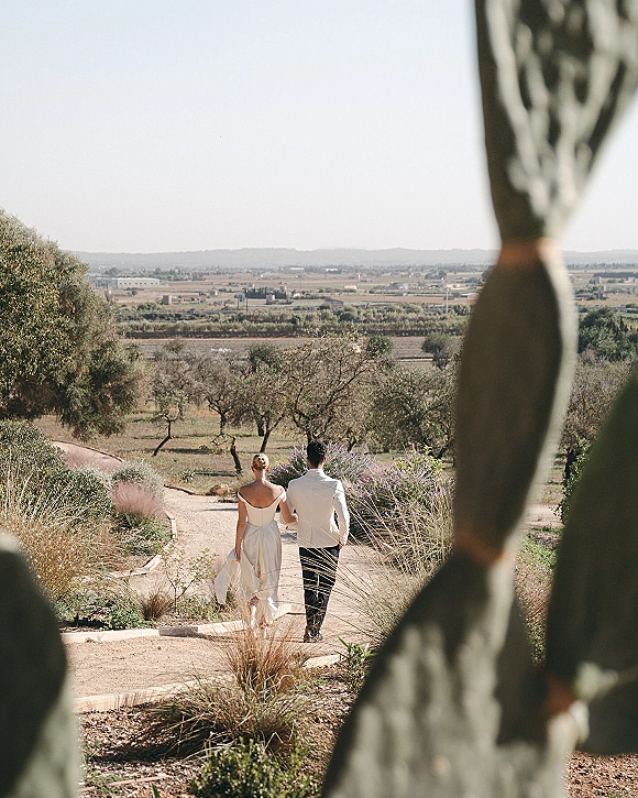 Couple portrait of newlyweds walking away on a desert path, bride in off-the-shoulder satin dress, groom in white jacket by cactus