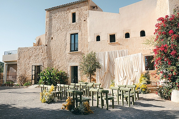 Ceremony setup for an outdoor wedding ceremony with green chairs in a semicircle, white draped arch and yellow aisle florals in a villa courtyard