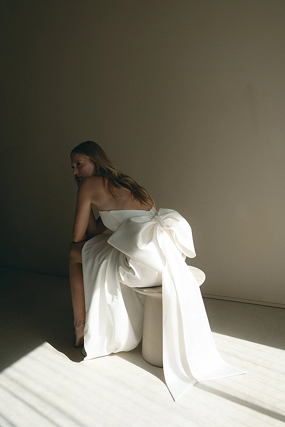 Bridal portrait of a bride in a strapless wedding dress, seated in side profile with an oversized bow and long tails in window light