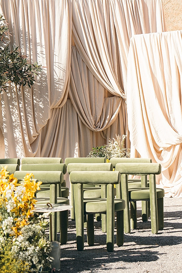 Ceremony setup with outdoor ceremony seating facing a draped fabric backdrop, green chairs, and floral greenery in a sunlit stucco courtyard