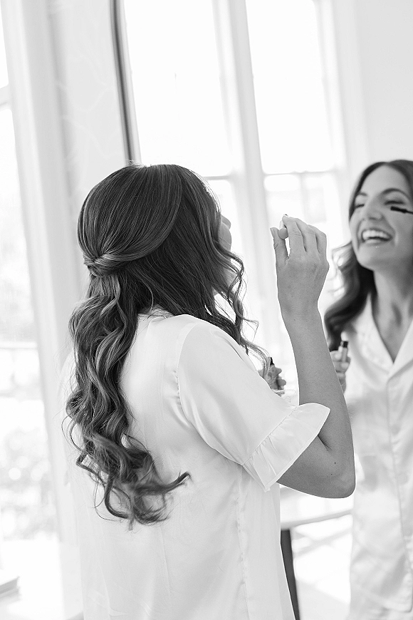 Bridal getting ready as the bride applying mascara in a satin pajama set, smiling in a mirror with soft window light in the room