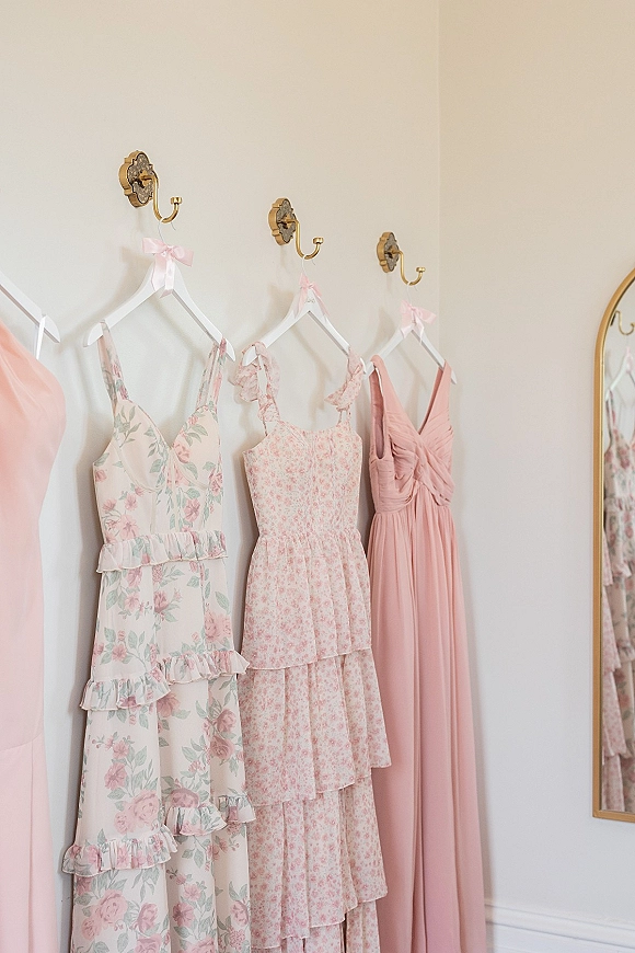 Bridesmaid dresses in blush tones hang from brass wall hooks with pink ribbon bows, including floral and tiered ruffle styles on a cream wall