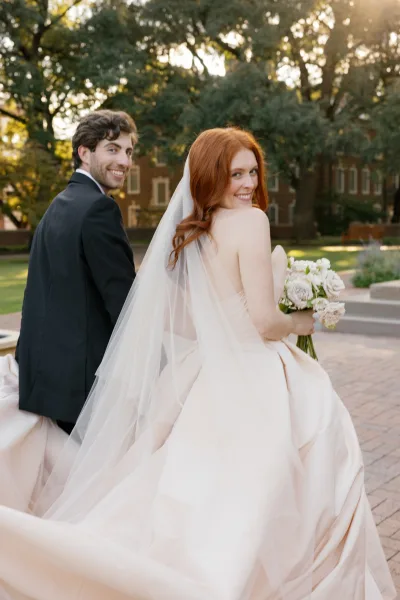Couple portrait of bride and groom looking back while walking down a brick walkway, her long veil and bouquet flowing in golden light